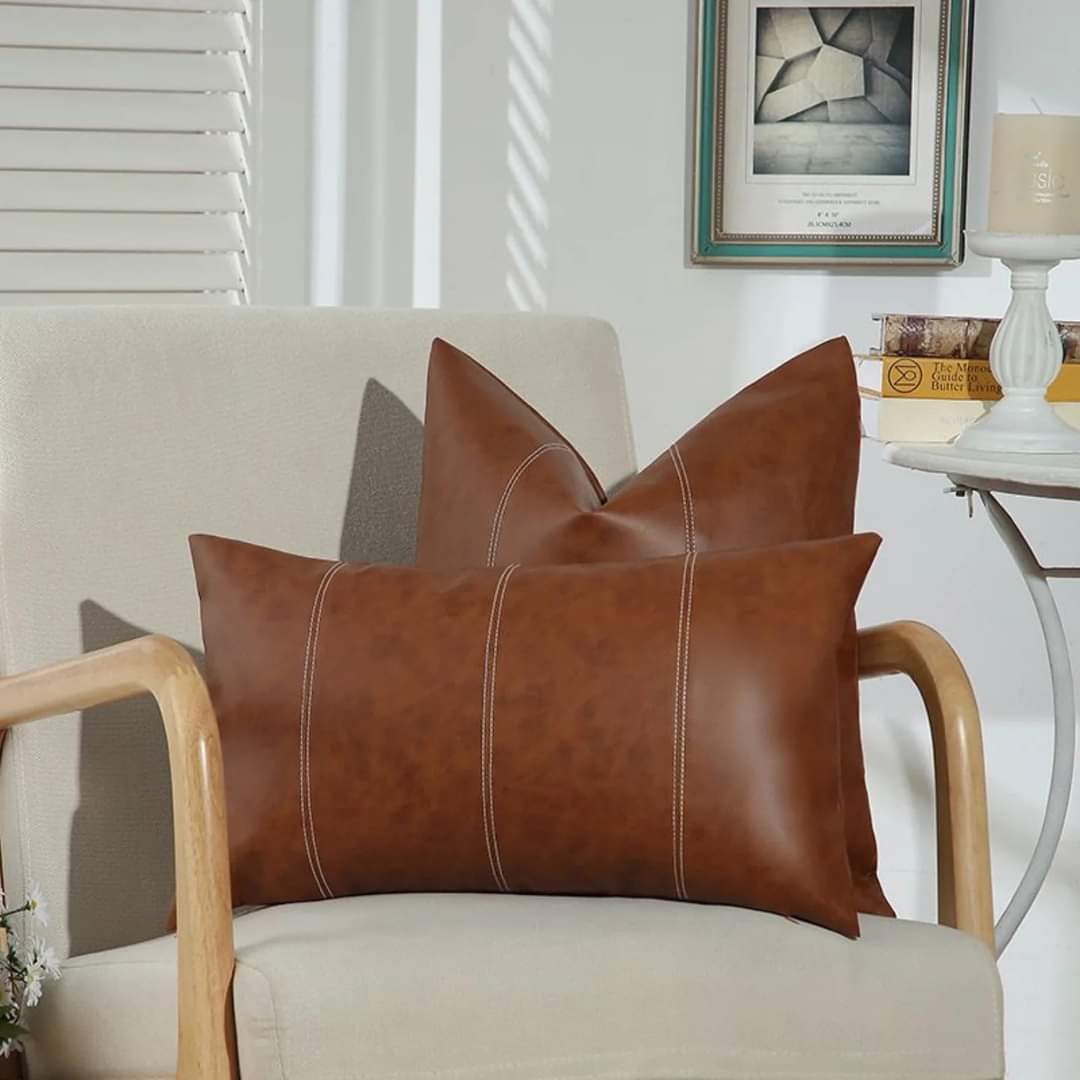Two brown faux leather rectangular throw pillow cases with white vertical stitching resting on a cream-colored wooden armchair in a brightly lit room.