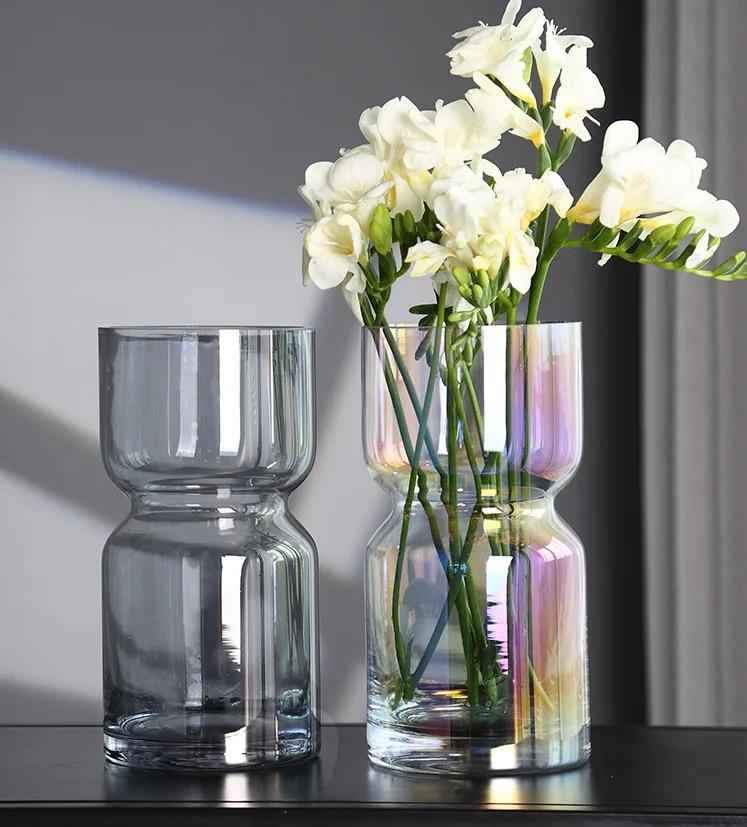 Two modern, hourglass shaped glass vases on a black surface; one in dark smoked grey and the other in iridescent glass