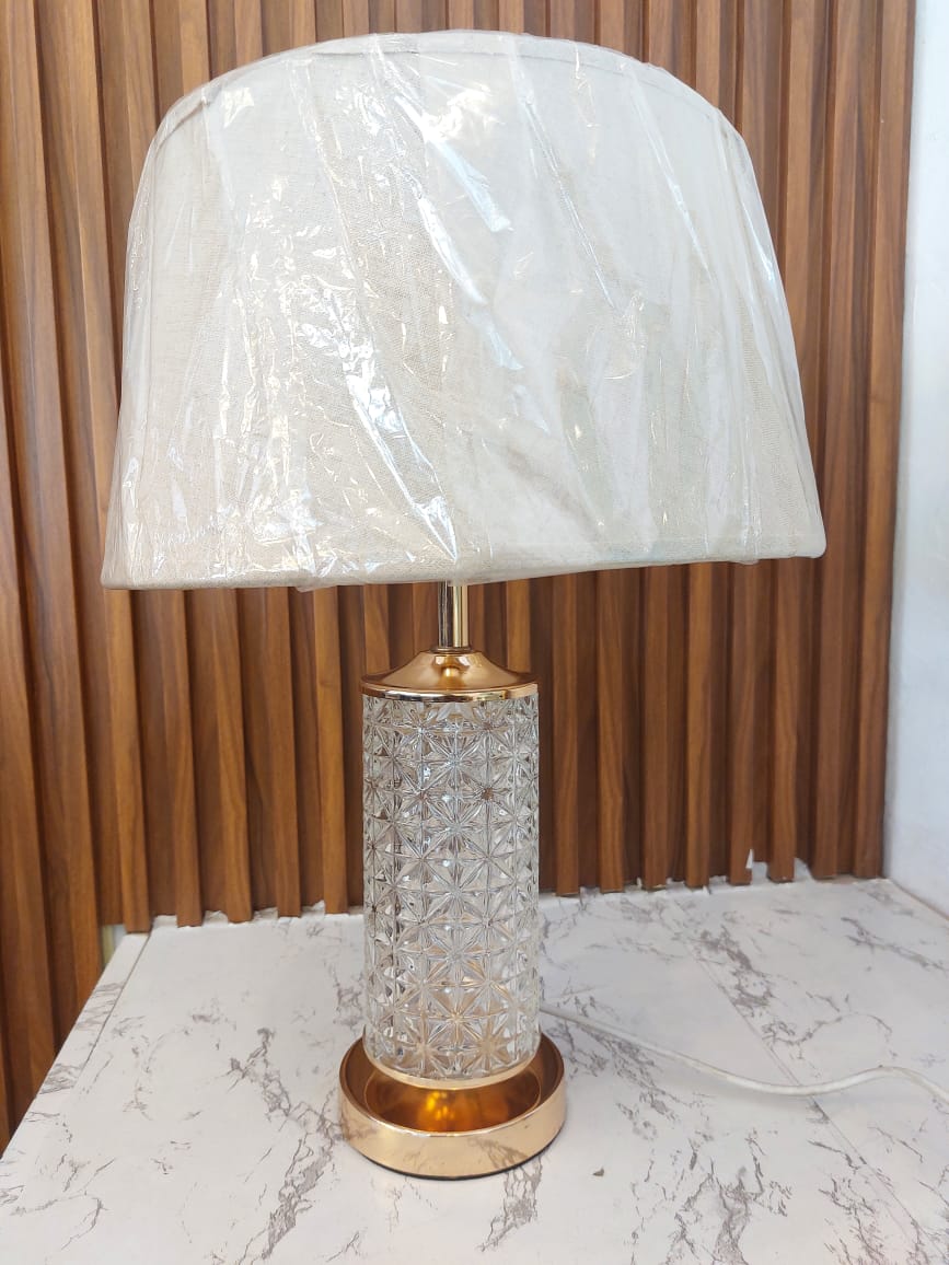 A table lamp featuring a textured geometric clear glass base with gold metal accents and an off white fabric shade.