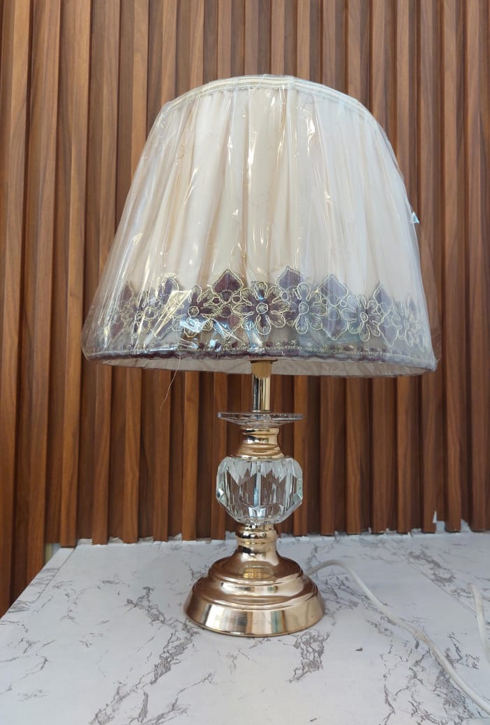 A table lamp featuring a cream pleated fabric shade with floral embroidery and a purple border on a gold base with a clear glass center piece.