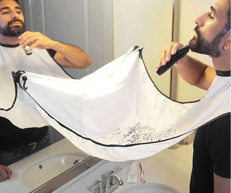 A man trimming his beard while wearing a white and black apron that attaches to the mirror to catch hair clippings over the sink.