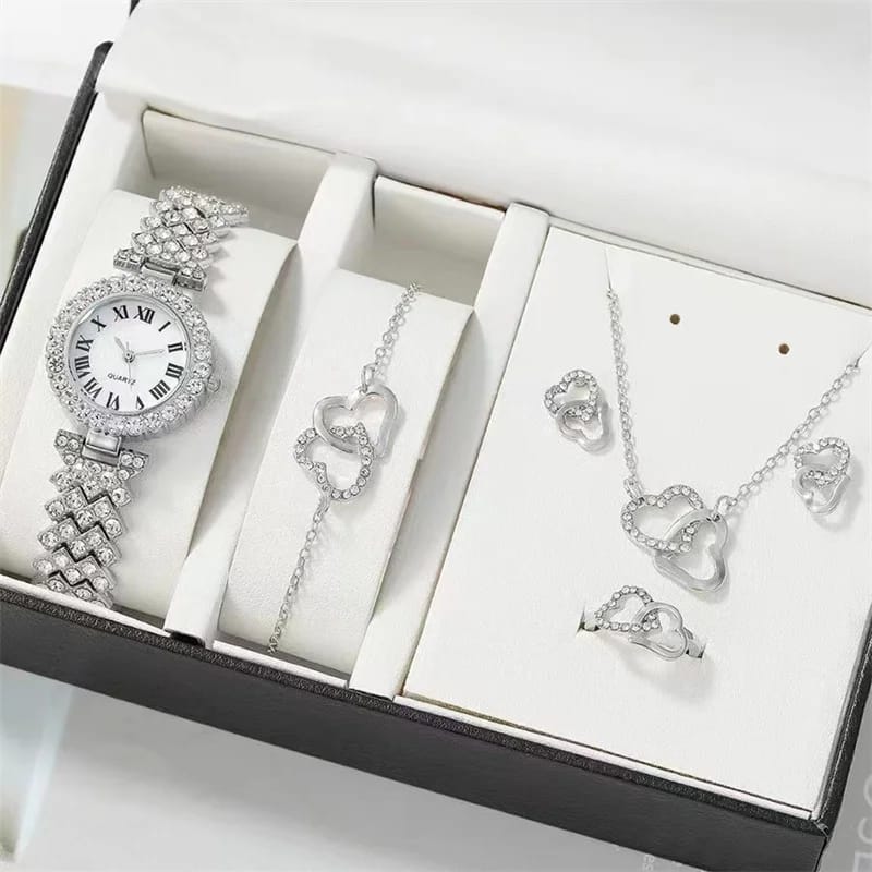 An open black and white gift box containing a silver-toned watch and a matching bracelet, necklace, earrings, and ring, all featuring a heart motif and clear stones.