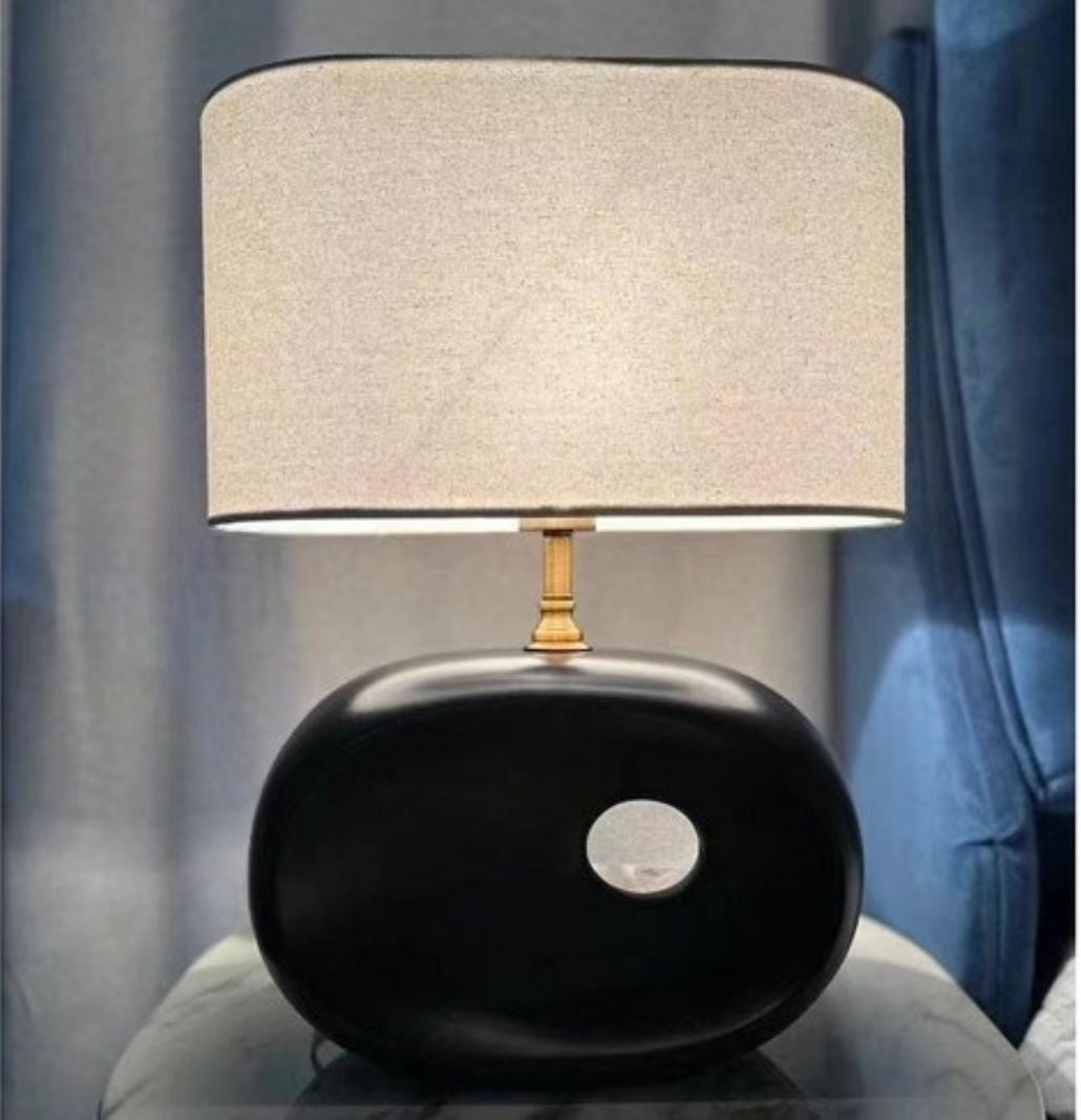 A table lamp with a black oval ceramic base featuring a circular cutout and a neutral fabric shade.
