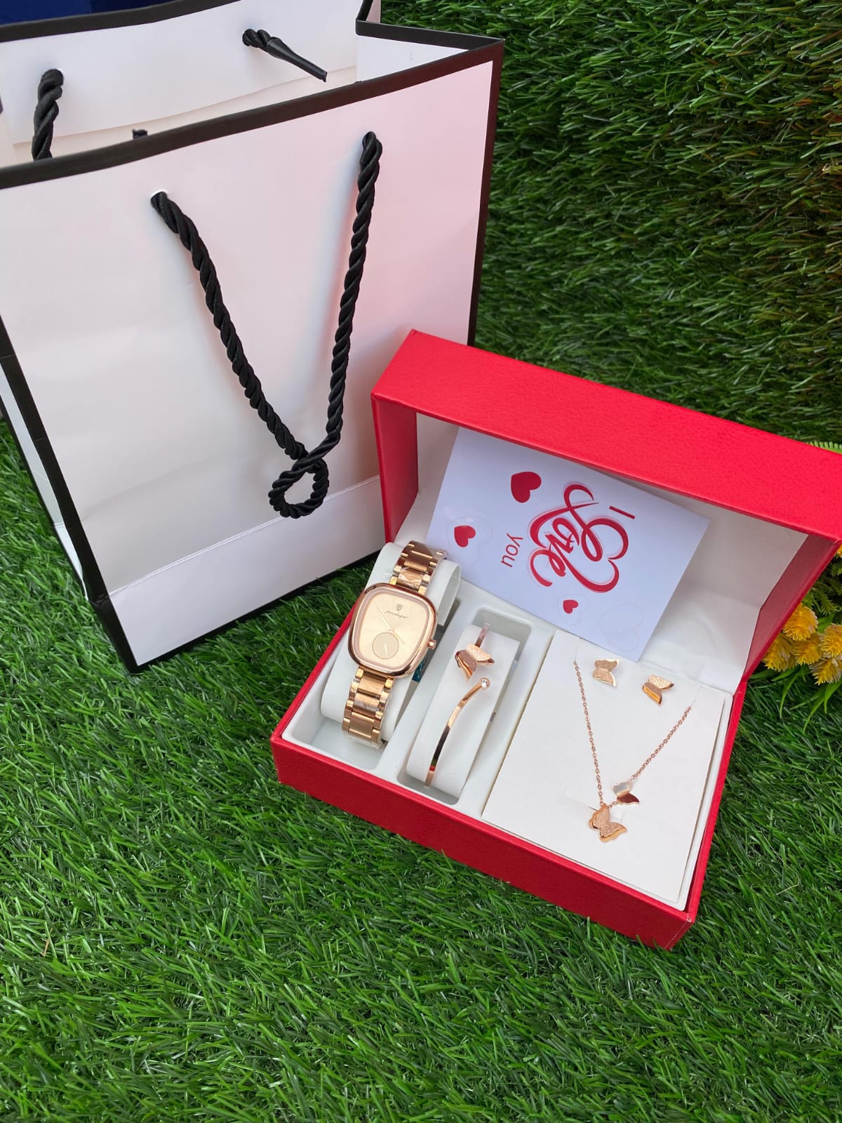 A red gift box containing a rose gold toned watch, matching butterfly necklace, bracelet, and earrings, sitting next to a white gift bag and a small "I Love You" card on a patch of green grass.