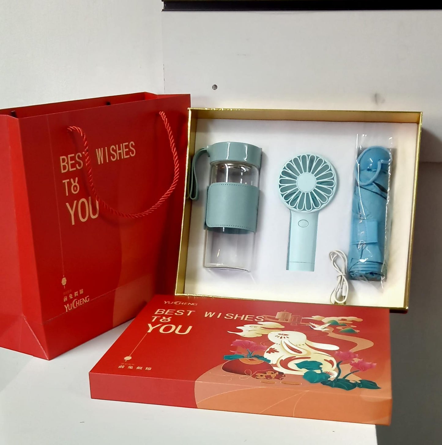 Gift set in a white box with a red gift bag, featuring a light blue mini fan, a glass bottle with a blue sleeve, and a compact blue umbrella.