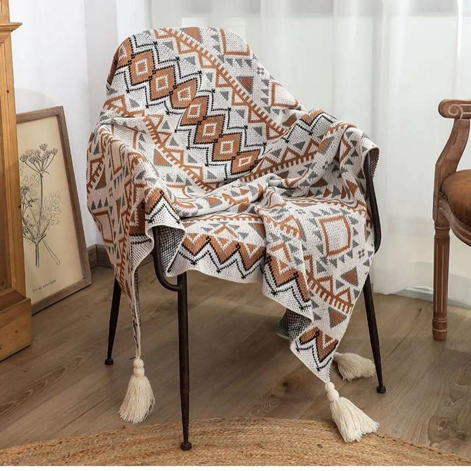 A bohemian knitted throw blanket with orange and grey tribal patterns and tassels