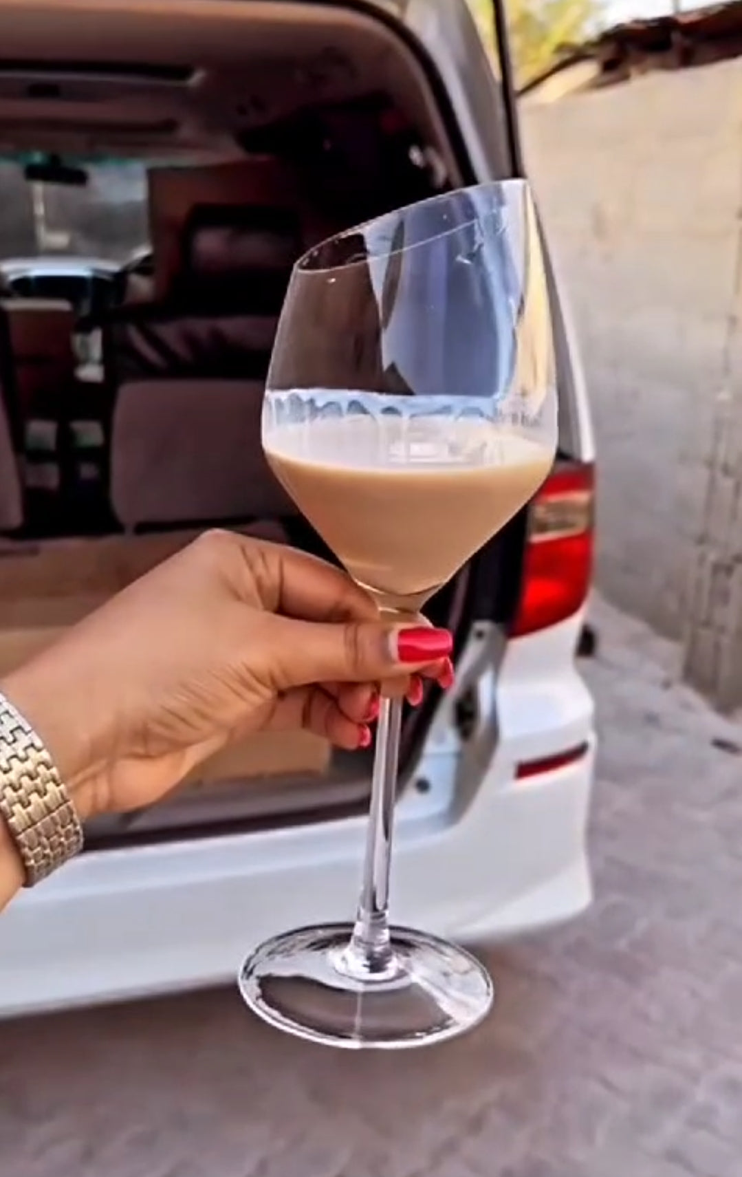 A hand holding a slanted large wine glass filled with a beige beverage, likely milk or a cocktail, with a vehicle in the background.