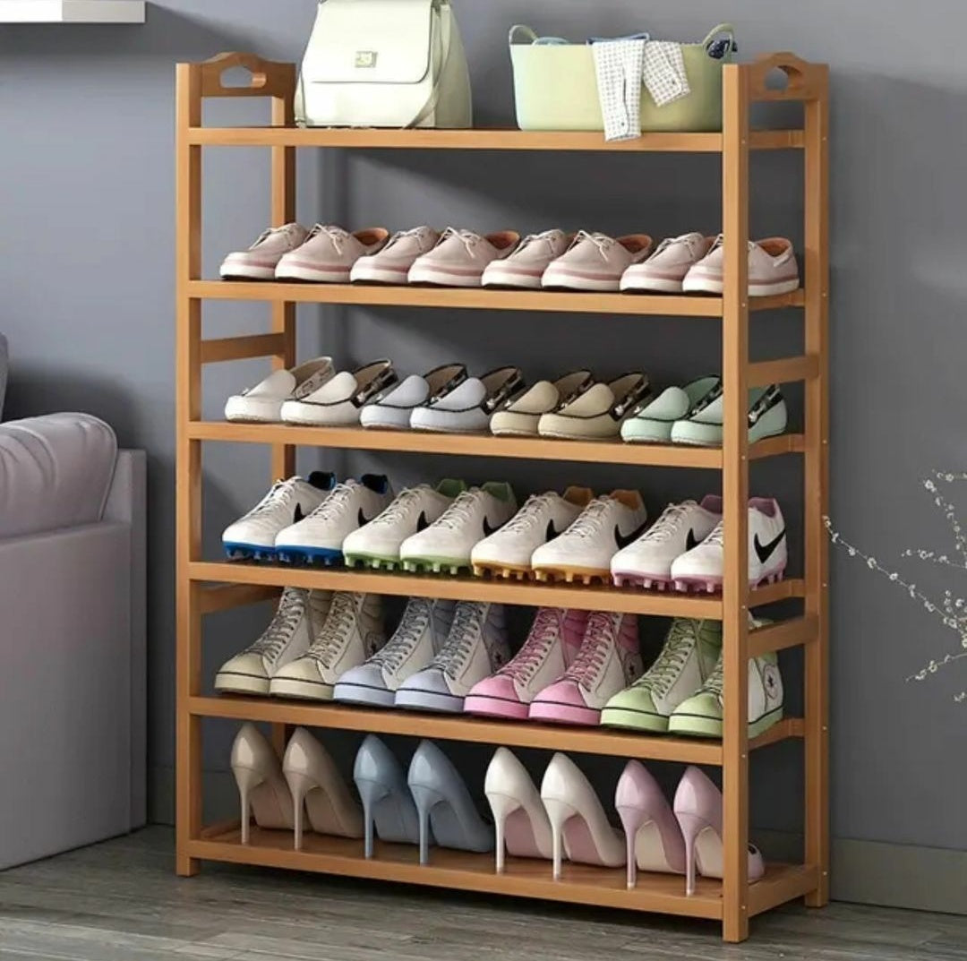A six tier, light brown bamboo shoe rack filled with various styles of footwear, including sneakers and high heels, standing against a grey wall in a living space.