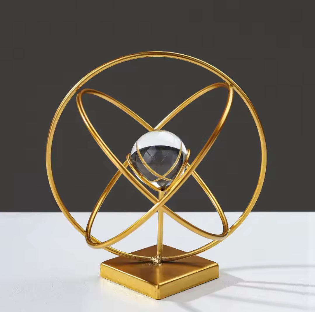 A gold metal orbital sculpture with a clear crystal center on a square base.