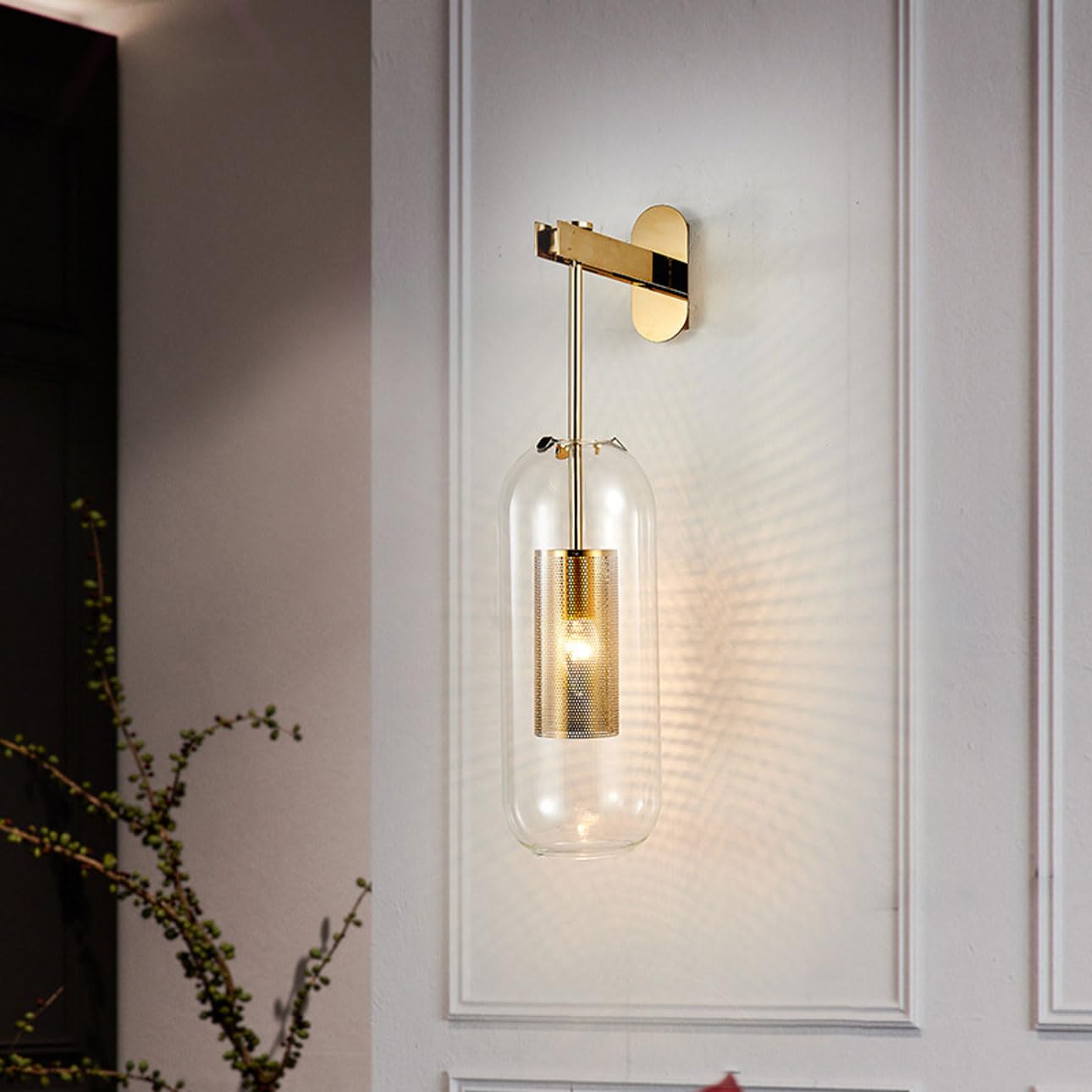 A gold wall lamp with a clear glass capsule shade and a mesh metal interior.