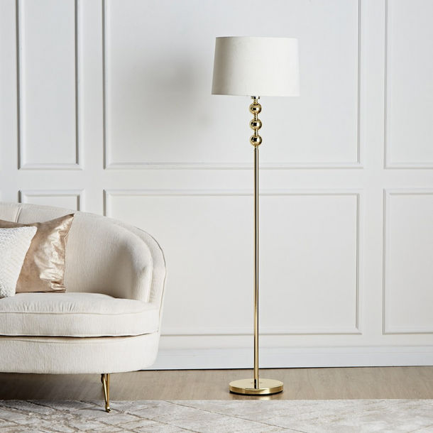 A tall gold floor lamp featuring three stacked metallic spheres on the upper neck and a white fabric drum shade.