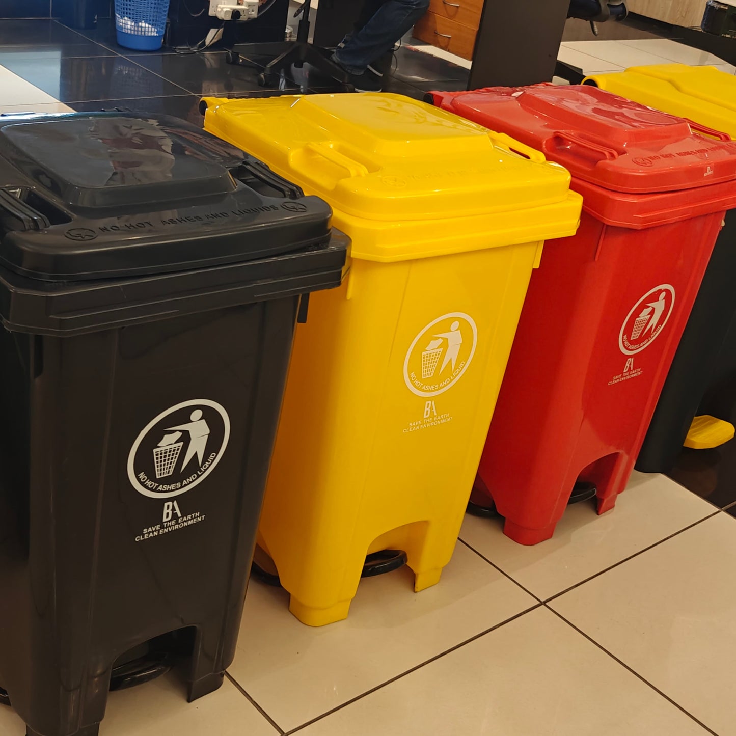 A row of tall, rectangular 100-liter plastic pedal bins in black, yellow, and red, each featuring a waste disposal graphic and text promoting a clean environment.