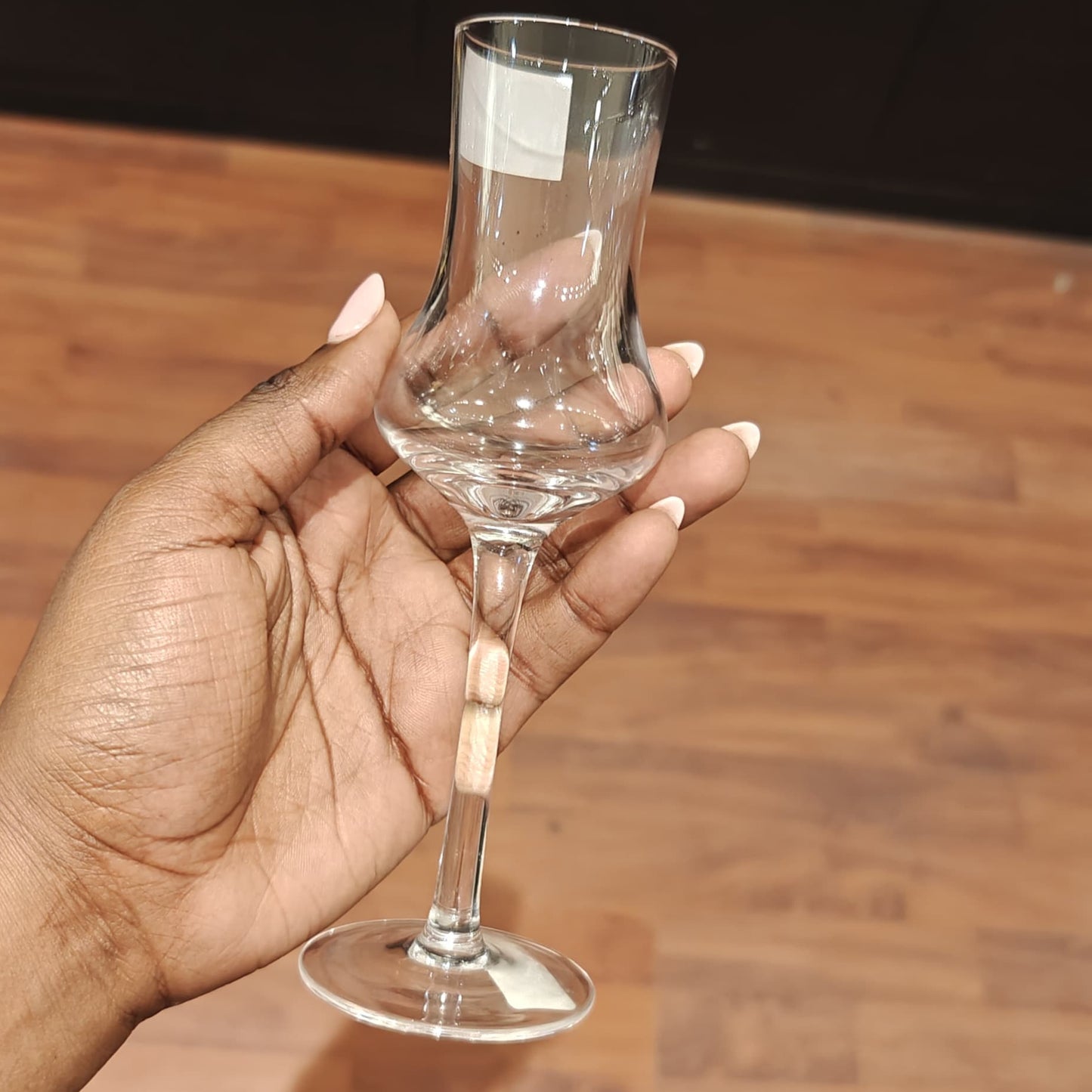 A hand with manicured nails holding a small, clear, tulip-shaped stemmed glass designed for serving grappa or cherry liqueur against a wooden floor background.