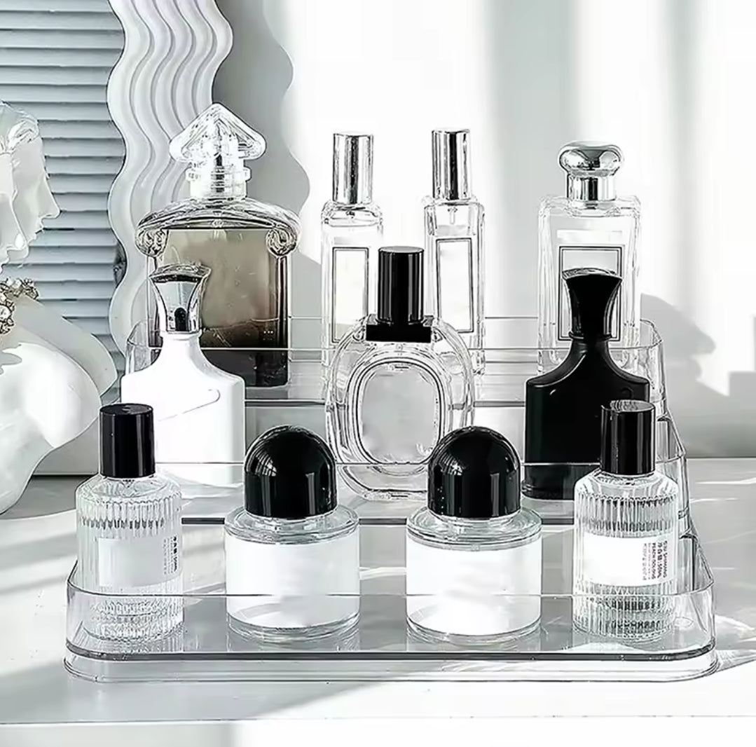 A tiered clear acrylic perfume stand holding several bottles of perfume and cologne on a white dresser.