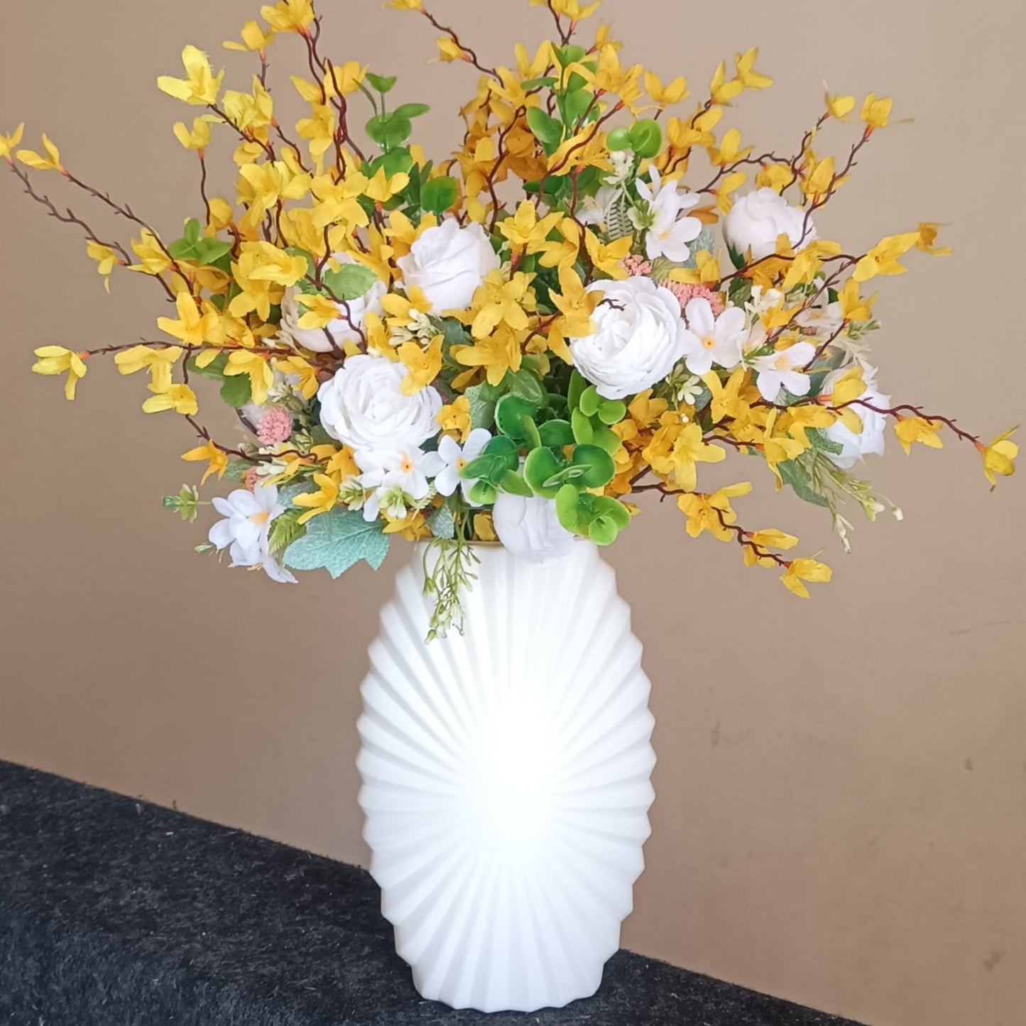 A large, vibrant artificial flower arrangement with yellow leaves, white roses, and small white filler flowers, set in a tall, white ribbed plastic vase.
