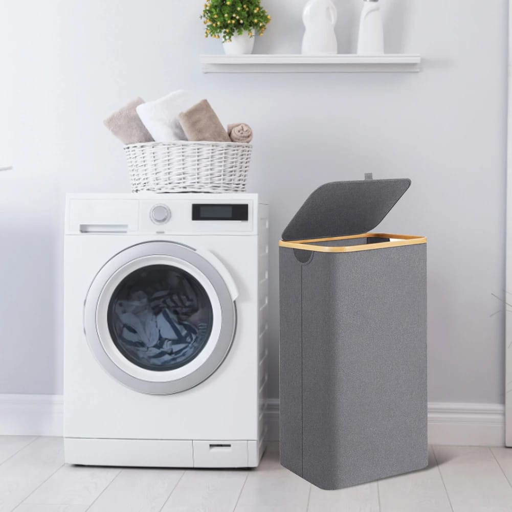 A tall, rectangular grey fabric laundry hamper with a wooden rim and an open lid, standing next to a white front loading washing machine.
