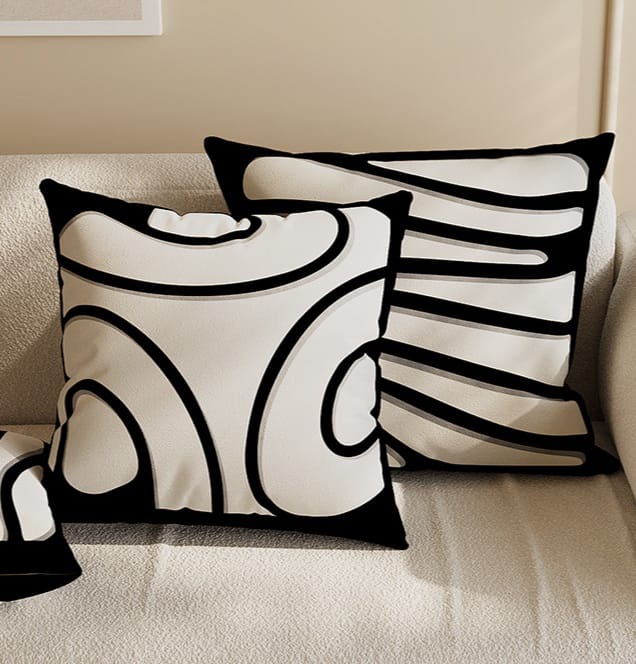 Two square throw pillow covers featuring bold, swirling black and white abstract graphic designs, displayed on a cream-colored textured sofa.