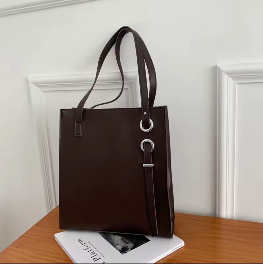 Dark brown structured vertical leather tote bag with long shoulder straps and decorative silver ring and buckle details.