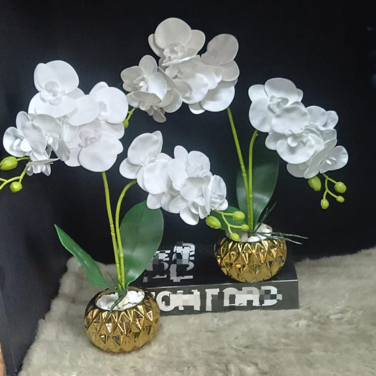 A small, round gold ceramic vase with a raised geometric pattern, containing stems of bright white artificial orchid flowers and green leaves.