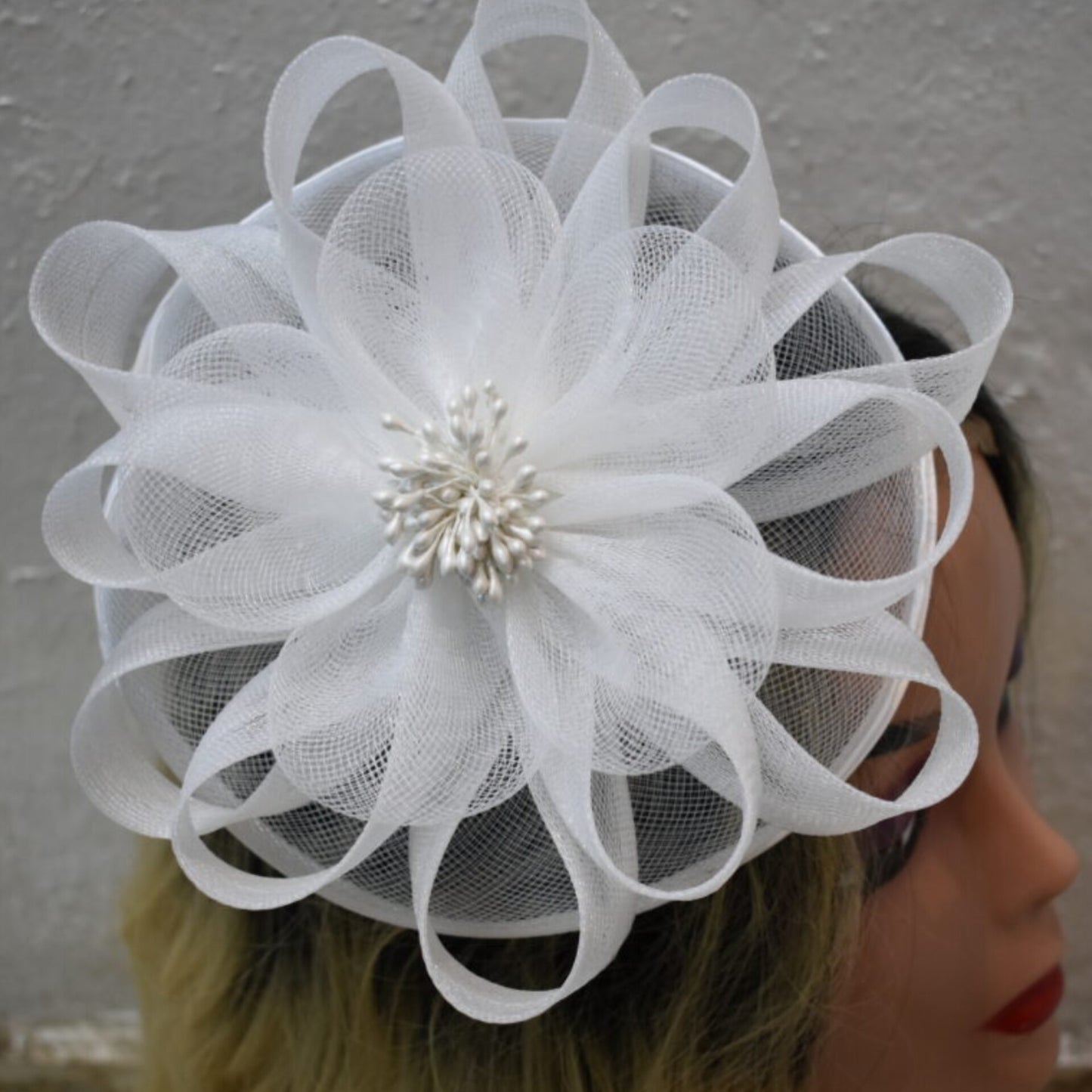 A white lady's fascinator made of looped organza fabric and pearl-like beads, modeled on a mannequin head with blonde hair against a textured white background.