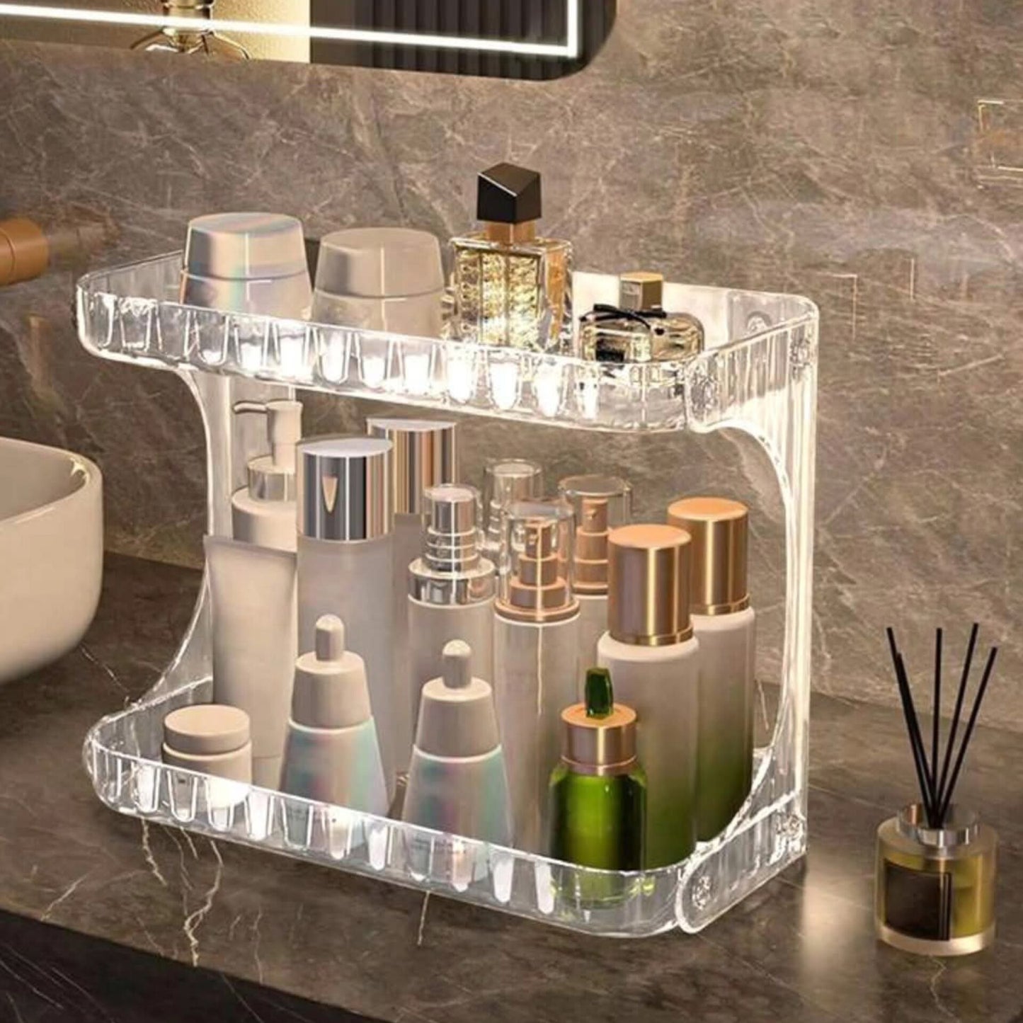Clear two layer acrylic shelving unit with textured sides, filled with various bottles of perfume and skincare products on a marbled bathroom counter.