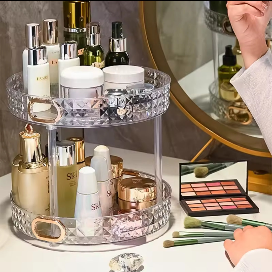 Clear two tier acrylic rotating cosmetic organizer with gold handles, filled with various skincare bottles and jars on a white marble vanity.