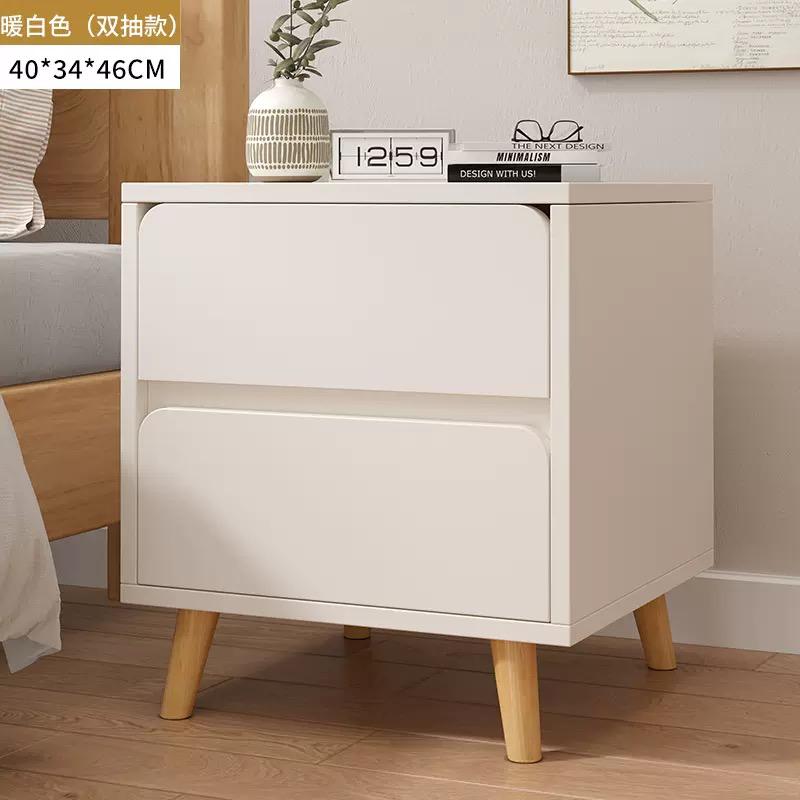 Warm white two drawer bedside table with minimalist fronts and natural wood legs, topped with a small vase and an alarm clock, next to a bed.
