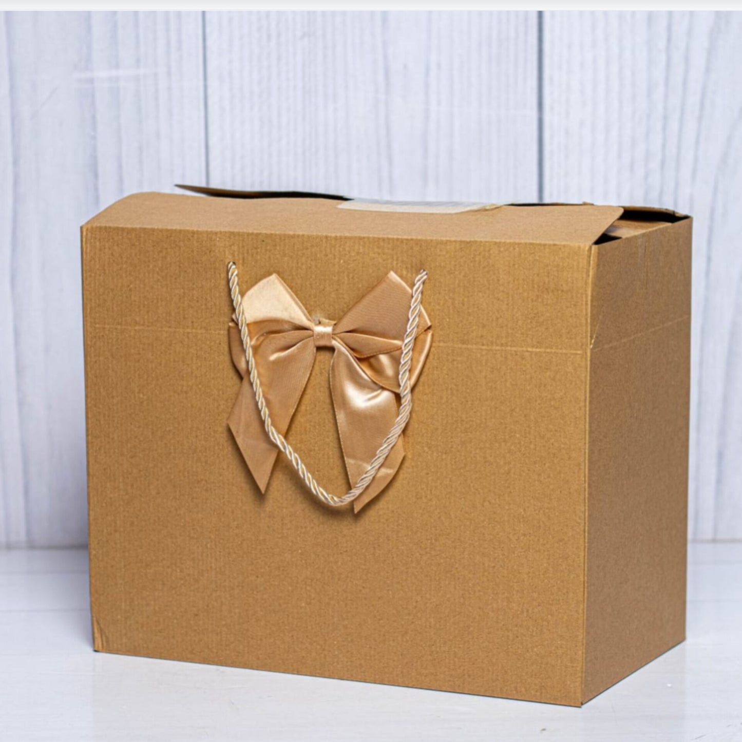 A rectangular, landscape oriented brown Kraft cardboard gift box with gold rope handles and a large decorative gold silk bow.