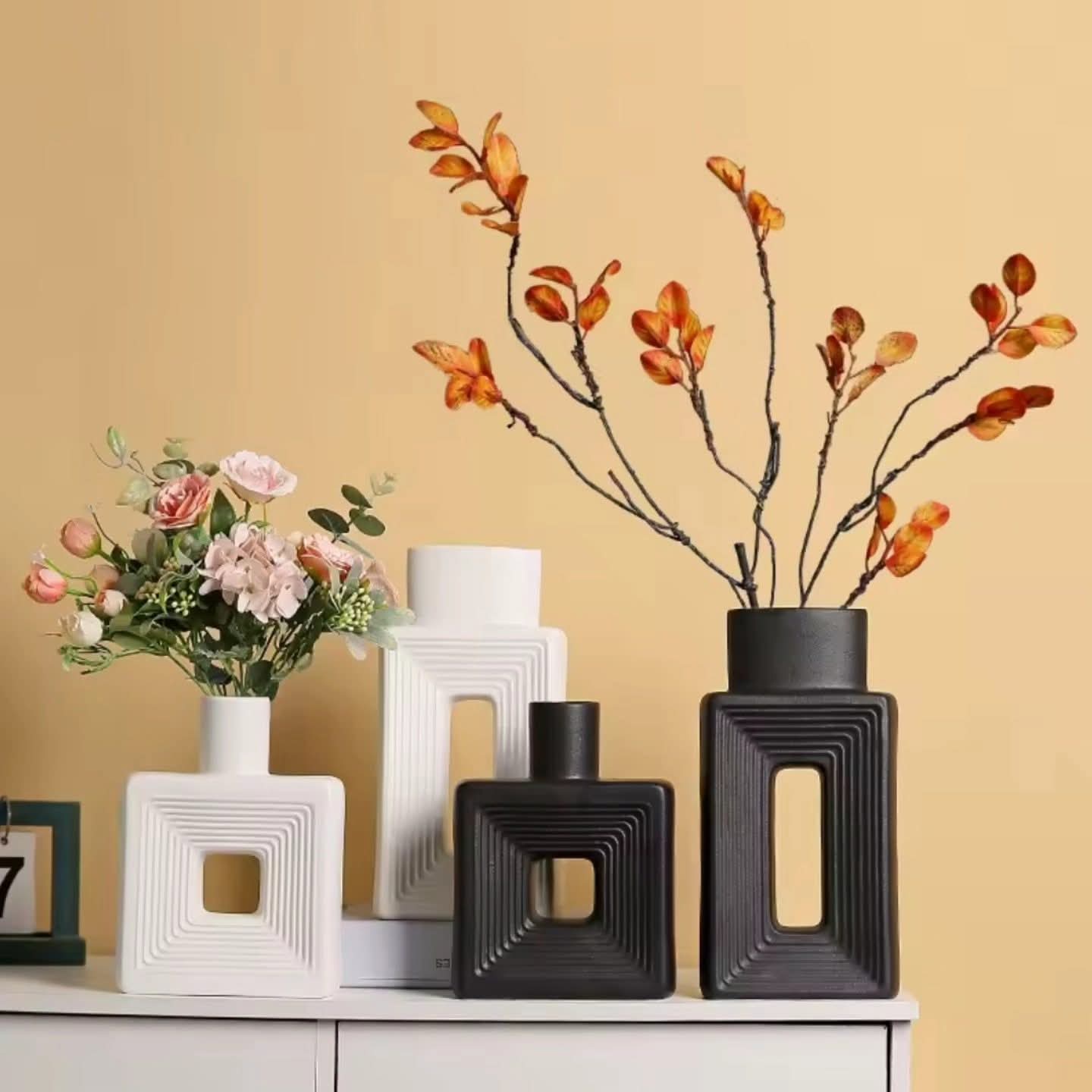 Two pairs of black and white square decorative vases of different heights, each with a cutout center, displaying pink roses and dried orange branches.