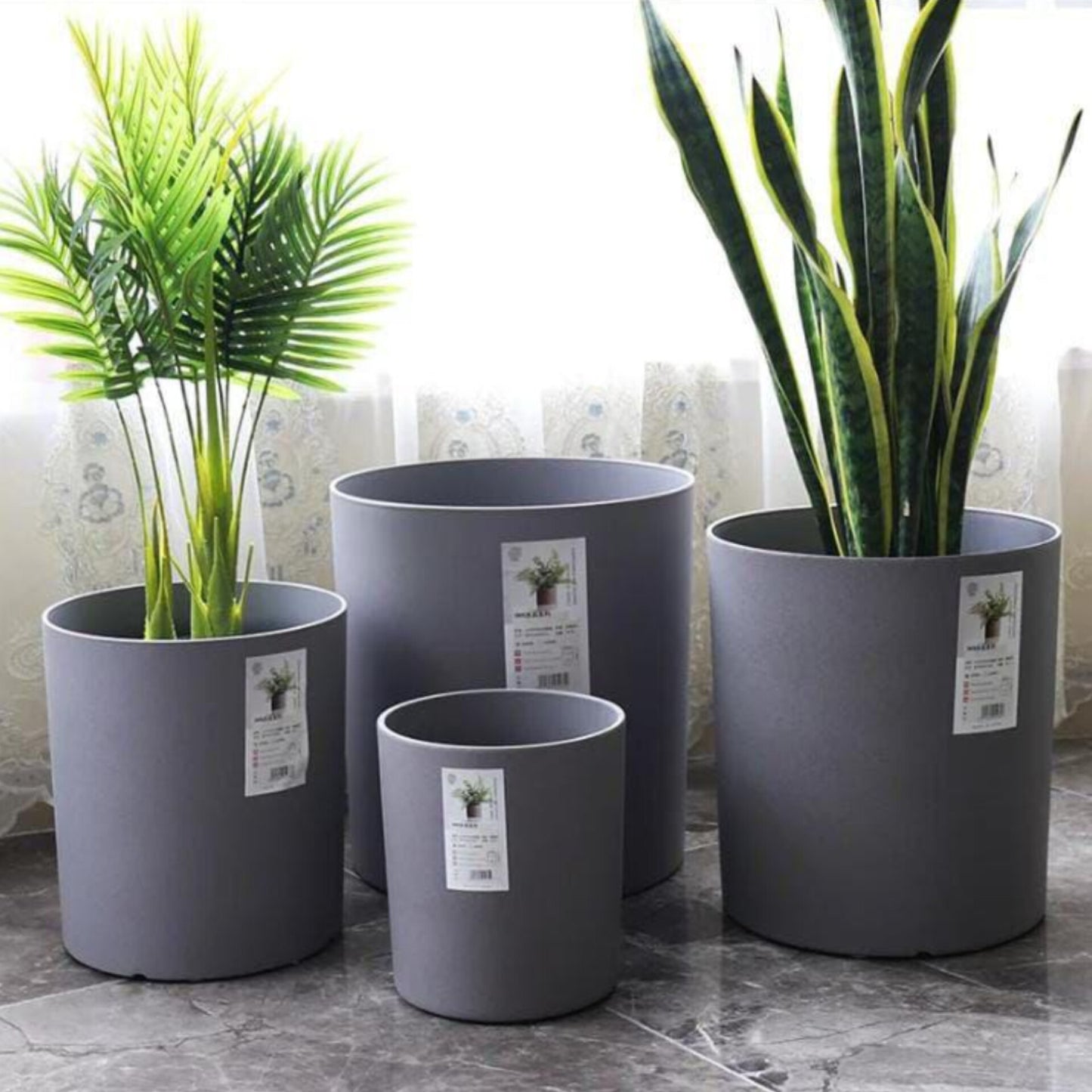 Four light grey cylindrical planters of varying heights displayed with indoor plants on a tile floor.