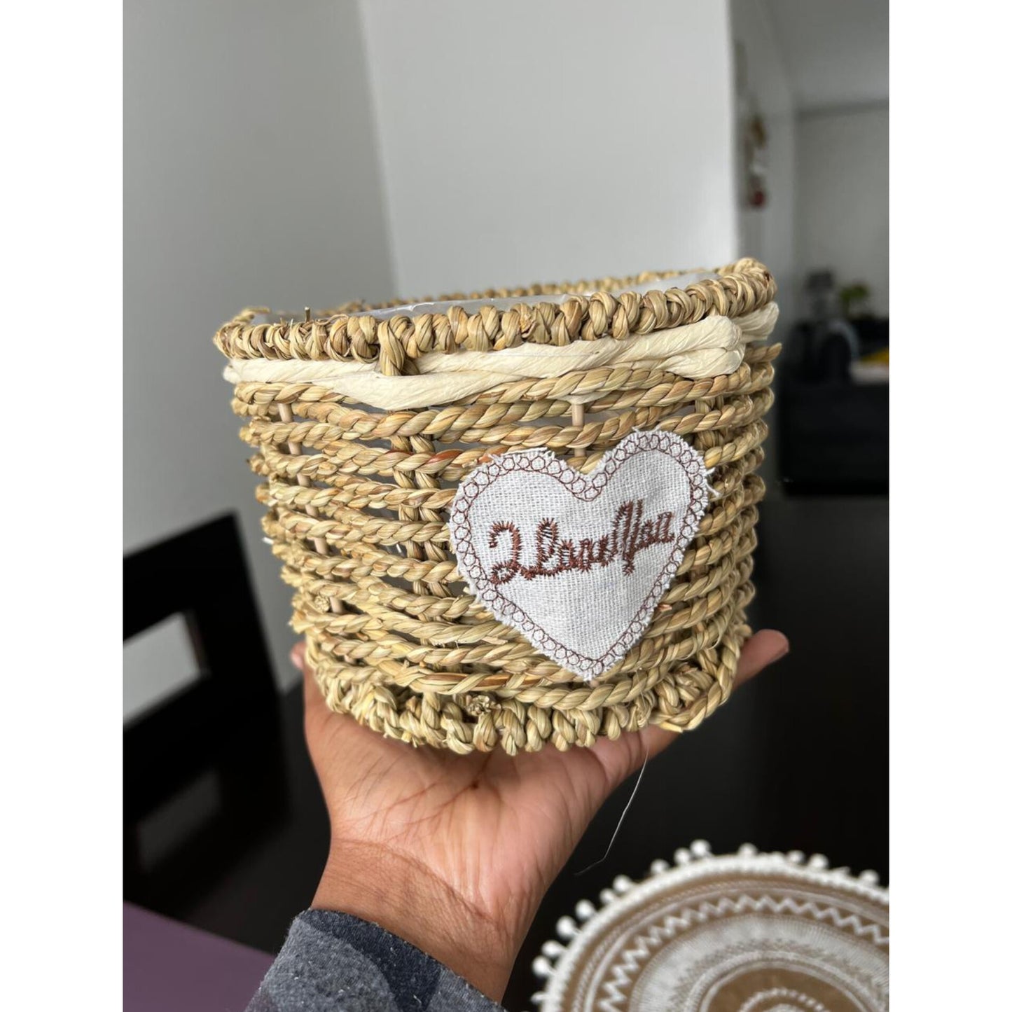A small, round basket hand-woven from natural straw-colored fibers, featuring a white fabric heart embroidered with "I Love You" on the front.