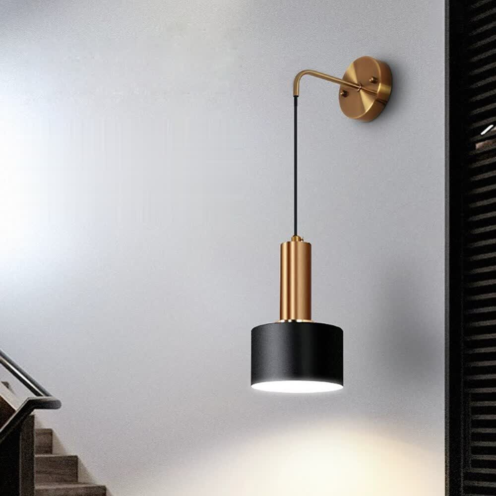A wall mounted lamp with a gold base and arm suspending a black cylindrical shade.