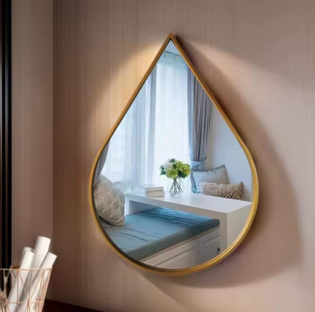 A modern teardrop shaped wall mirror with a thin gold frame reflecting a bright window area and flower arrangement on a built-in bench.