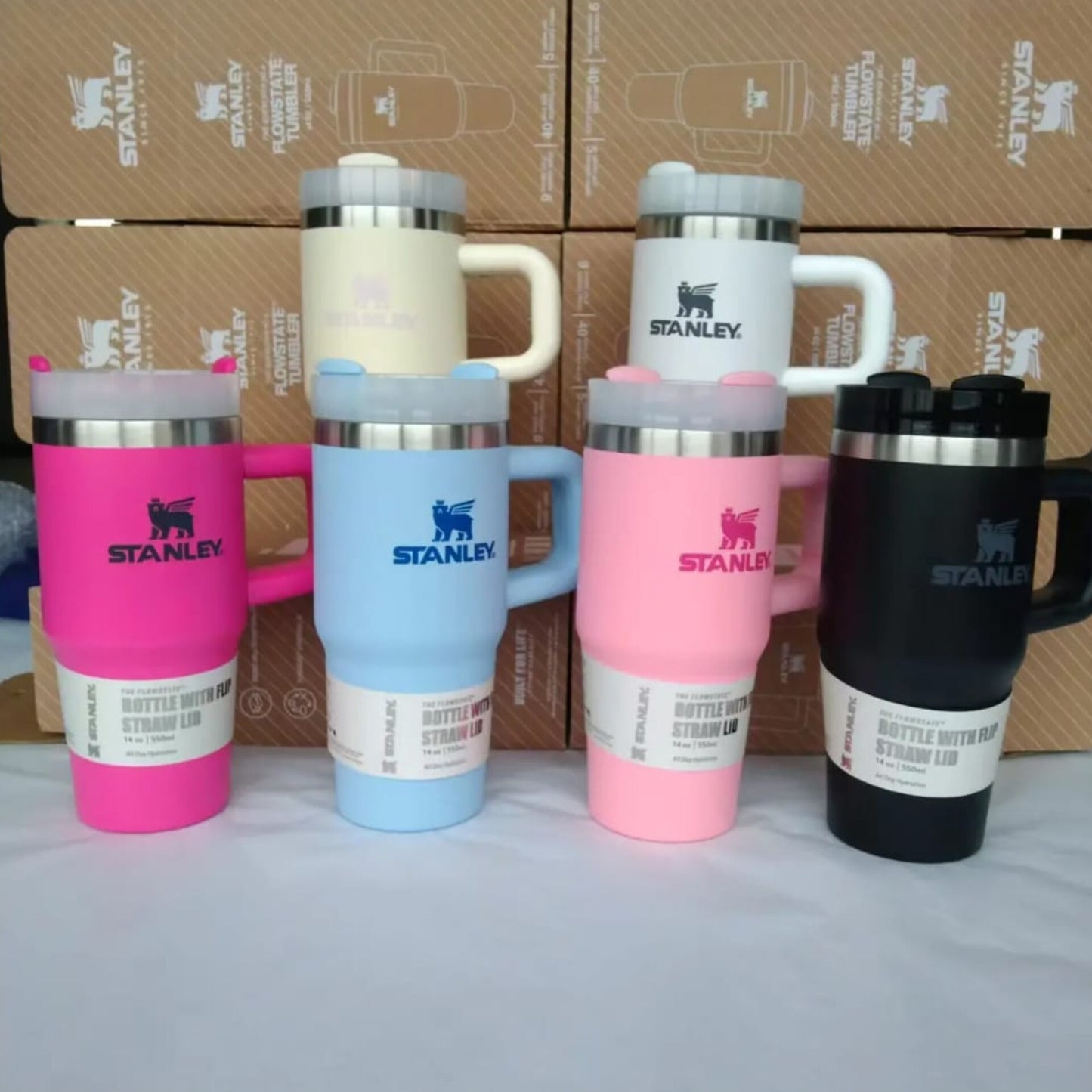A row of colorful Mini Stanley-style vacuum insulated mugs in pink, blue, black, and cream with handles and straw lids.
