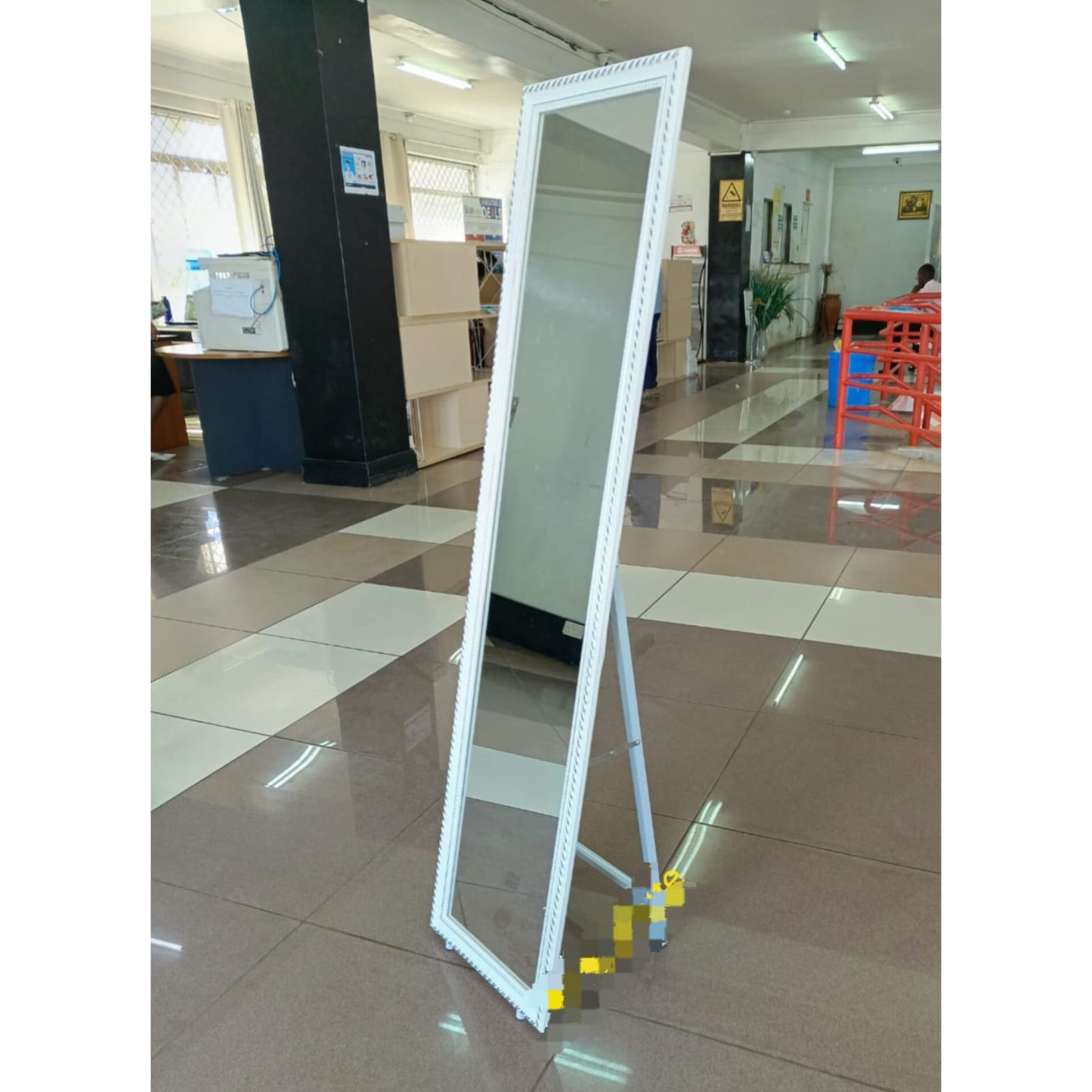 A tall, rectangular full body length mirror with a white decorative frame, supported by a metal A-frame stand, standing on a tiled floor in a retail space.