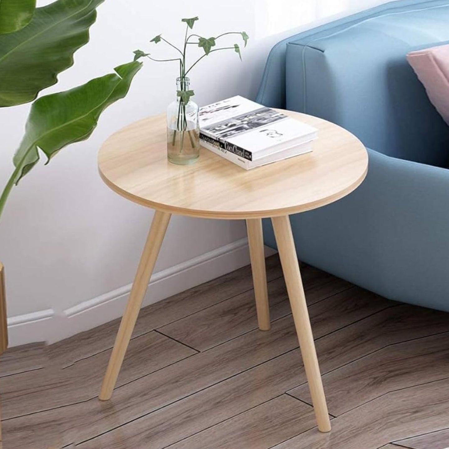 A simple light brown wood round table with three angled legs, holding a book and a clear vase with a sprig of greenery, placed next to a blue sofa.