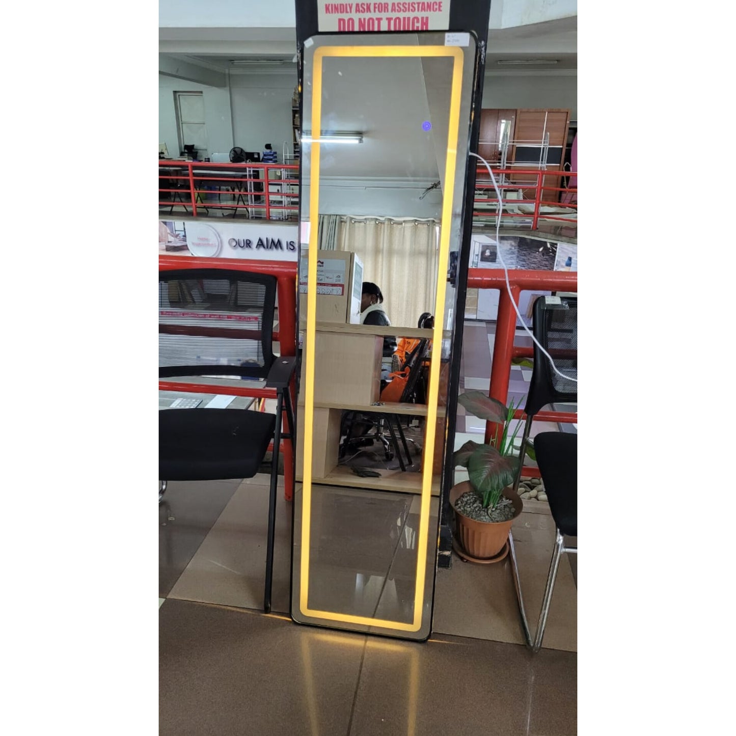A tall, rectangular full body length mirror with a warm yellow LED light strip outlining the frame, standing on a floor in an indoor retail space.