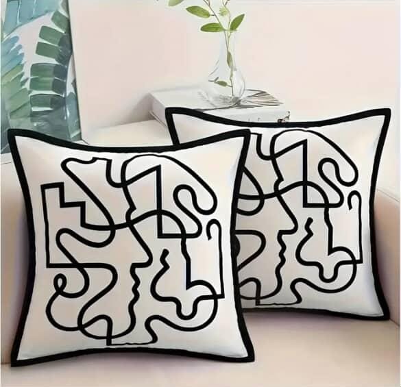 Two white square throw pillow covers with abstract black line art, featuring subtle profile faces, displayed on a light-colored sofa.