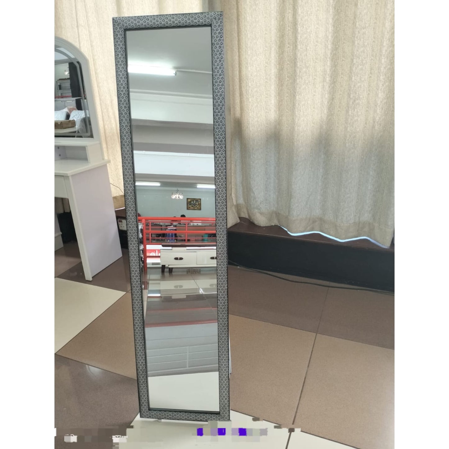 A tall, rectangular full body length mirror with a dark patterned frame, standing upright on a floor in an indoor space with sheer curtains in the background.