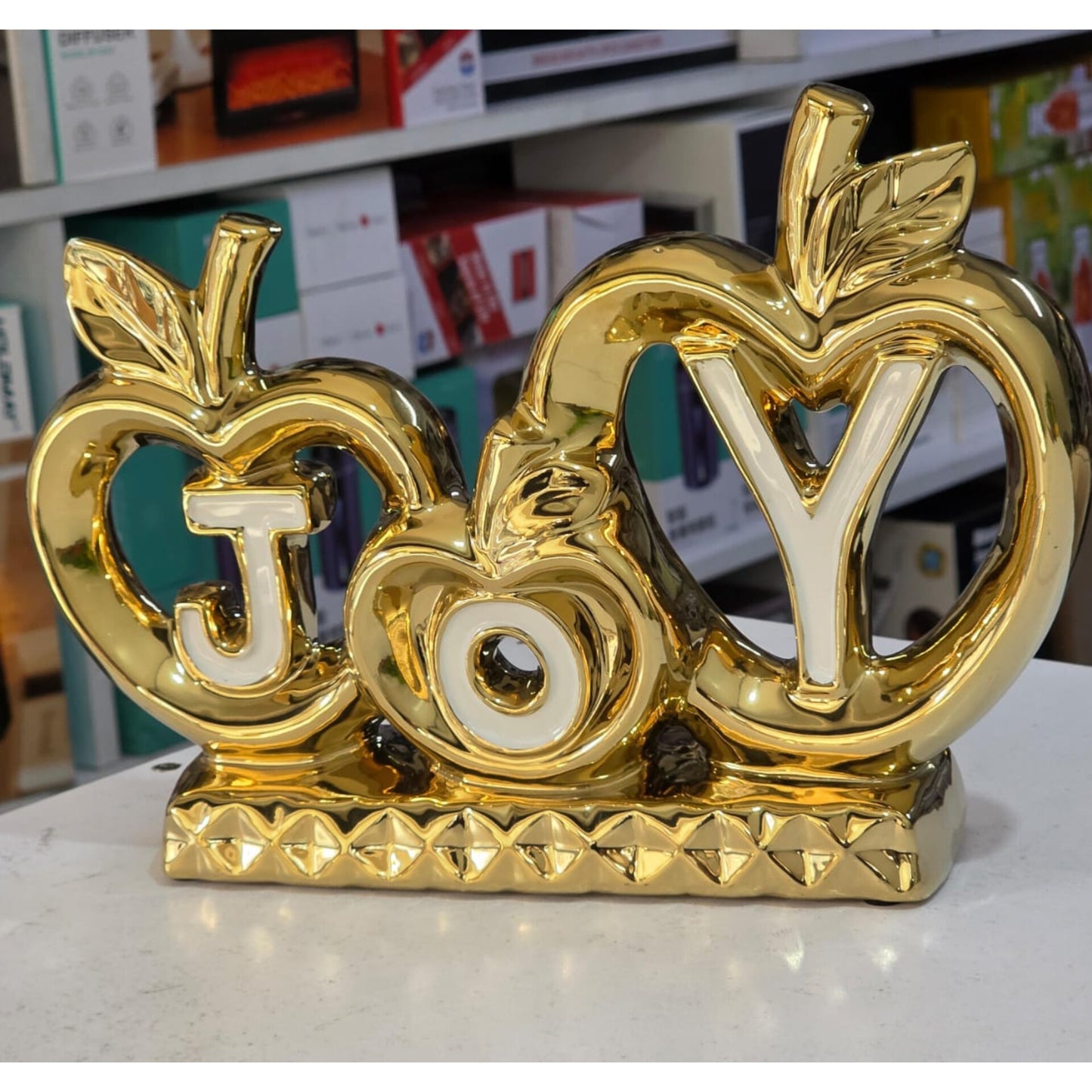 A gold ceramic tabletop ornament depicting two interlocking apple shapes with the word "JOY" cut out and filled in white, standing on a textured gold base.