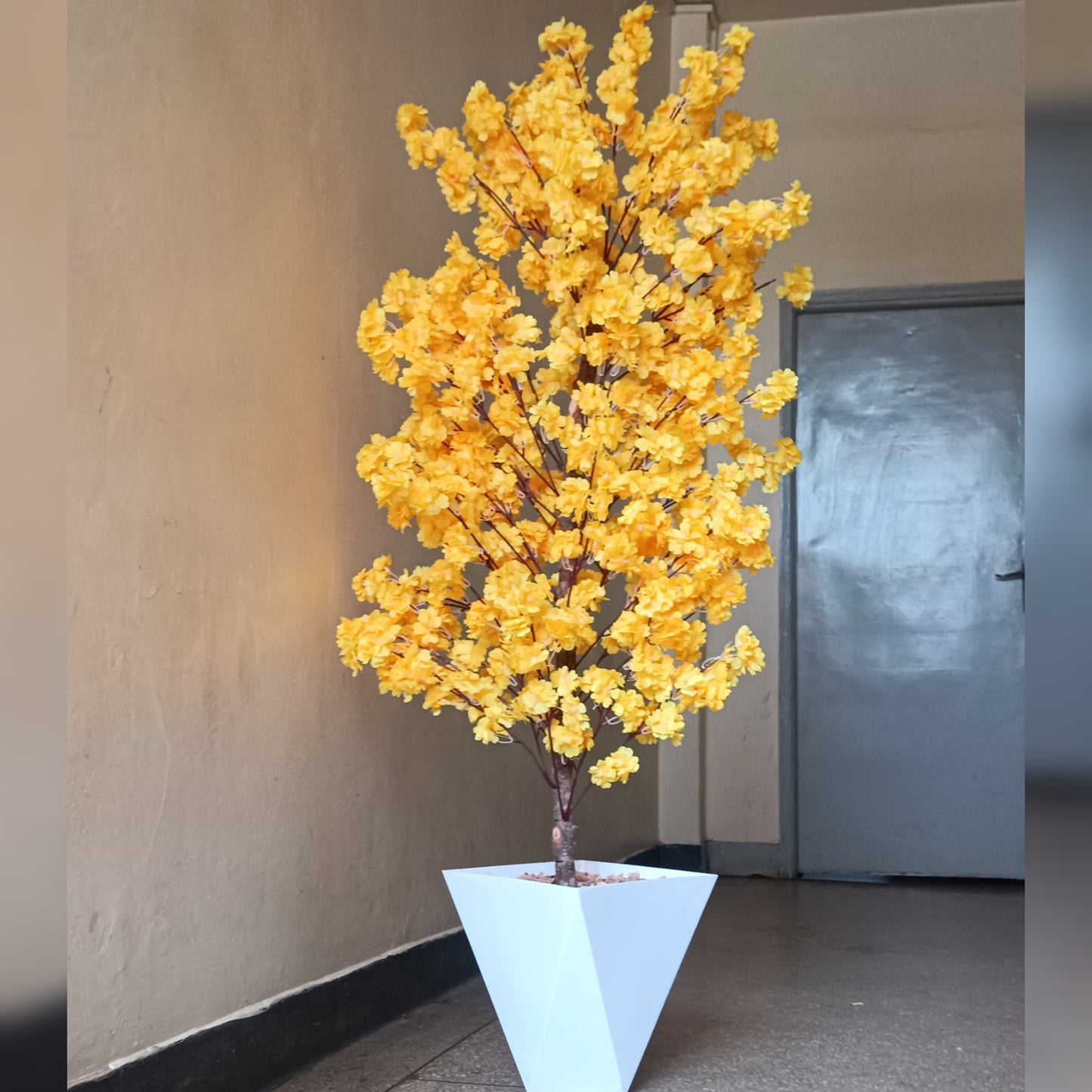 A tall artificial cherry blossom tree with vibrant yellow flowers in a white geometric vase.