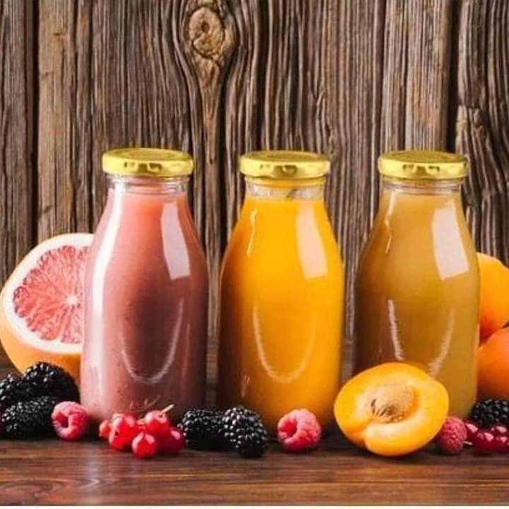 Three glass bottles filled with pink and orange juice standing on a wooden surface next to various fruits like grapefruit, berries, and a sliced apricot, all topped with shiny golden lids.