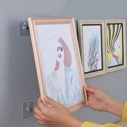 Two hands demonstrate placing a framed picture onto a clear, no drilling adhesive wall hook on a gray wall, with two other frames already hung nearby.