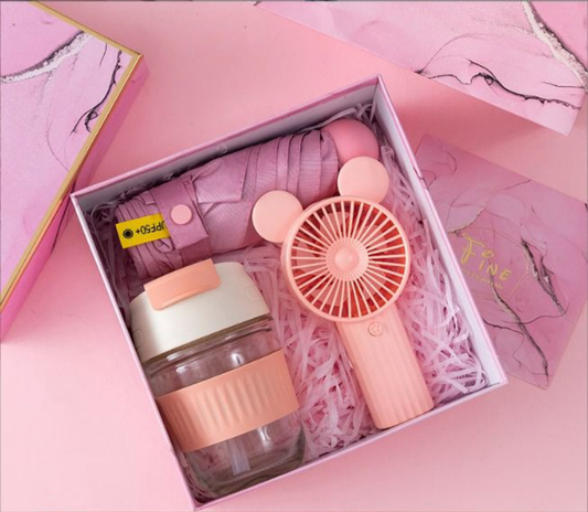 An open pink gift box containing a glass water bottle with a silicone band, a folded pink umbrella, and a pink handheld mini fan on shredded pink paper.