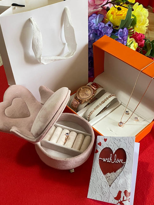 A gift box containing a rose gold-toned women's watch, necklace, and earrings, with a heart-shaped pink jewelry box, gift card, and pink gift bag.