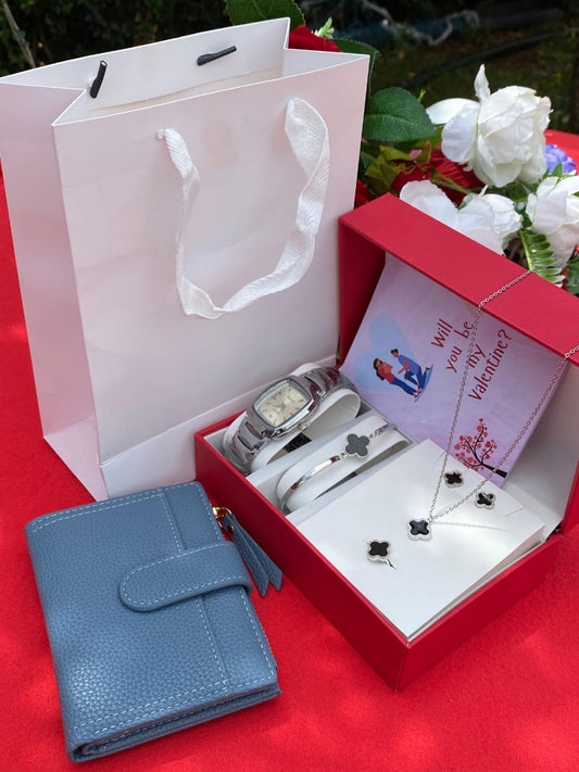 A red gift box open to show a silver watch and clover-themed necklace, earrings, and bracelet, next to a blue ladies' wallet and a white gift bag.