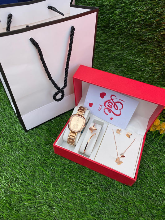 A red gift box containing a rose gold toned watch, matching butterfly necklace, bracelet, and earrings, sitting next to a white gift bag and a small "I Love You" card on a patch of green grass.