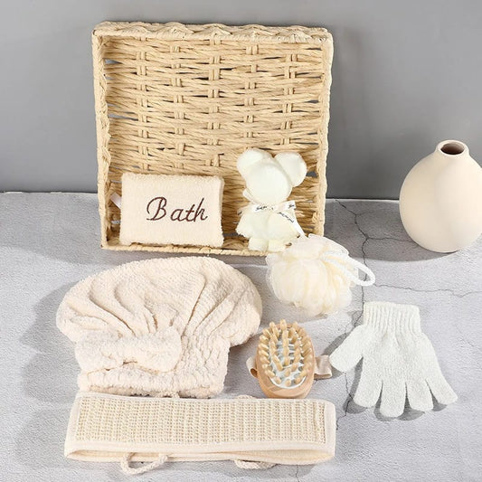 A woven basket containing various neutral-colored bath and spa accessories, including a back scrubber, exfoliating glove, bath pouf, hair towel, a scrub brush, and a bear-shaped soap.