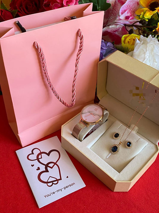 Rose gold-toned watch and jewelry set with blue stones, including a necklace and earrings, packaged in a beige box next to a pink gift bag and a card.