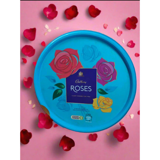 A large, round, bright blue Cadbury Roses chocolate tin decorated with colorful rose illustrations, surrounded by pink and red rose petals on a pink background.